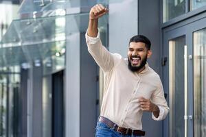 indio joven hombre en pie cerca un oficina edificio y alegría en éxito levantamiento su mano arriba y gritos con felicidad, él tiene un trabajo, un promoción, un negocio acuerdo foto