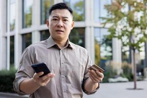 Worried Asian male office worker looking sad at camera, standing outside office, holding phone and credit card. photo