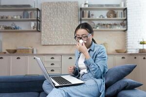 Work from home. Hospital. A young Asian woman freelancer fell ill. Works from home on the sofa at the laptop. He feels bad. He has a runny nose, wipes his nose with a napkin. photo