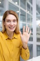 vertical disparo, hembra empleado mirando a teléfono cámara ondulación y sonriente, mujer de negocios hablando remotamente utilizando teléfono inteligente aplicación dentro oficina a lugar de trabajo. foto