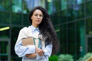 Portrait of confident Indian female student standing in front of office outside holding notebook looking at camera, young office worker with curly dark hair wearing glasses. photo