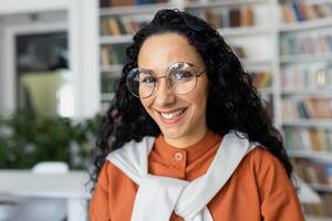 Portrait of young beautiful female student inside university academic library, Hispanic woman with curly hair and glasses close up smiling and looking at camera. photo