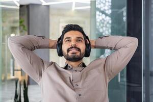 Close-up photo of a young Indian man in headphones who works in the office, sits thoughtfully at the table and rests with his hands behind his head.