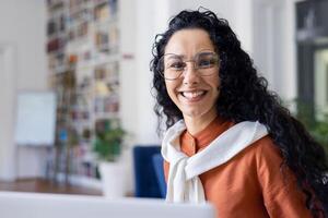 Side view of brunette female in round eyewear and white scarf over orange blouse smiling at camera. Friendly latina woman welcoming joining book club at college with satisfied facial expression. photo