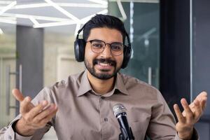 cámara web vista, hombre en auriculares mirando a cámara, sonriente, utilizando profesional micrófono a grabar podcast formación en línea curso, empresario entrenador dentro oficina. foto