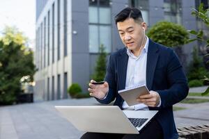 Asian young businessman sitting near office building on a bench wearing headphones, talking on a call through a laptop and making notes in a notebook. photo