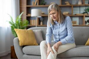 Pensioner woman sitting on grey couch and holding knee while bending over with grimace of pain. Suffering lady holding leg with inflammation related to ageing process and joint degeneration. photo