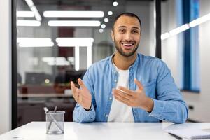 Photo of a smiling young hispanic man in a denim shirt sitting at a desk in a happy office and talking to the camera via call.
