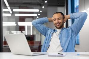 Smiling and relaxed young hispanic man sitting in the office at the desk, resting his hands behind his head and closing his eyes. photo