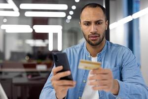 A man is looking at his phone while holding a credit card. He seems to be checking his balance or making a purchase photo