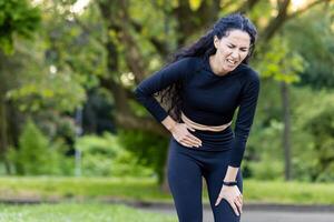 A Hispanic woman dressed in sportswear clutches her stomach in pain while jogging in a park, expressing discomfort and pain on her face. photo