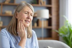 An elderly Caucasian woman sits at home, touching her cheek and grimacing in pain. She appears to be suffering from a severe toothache or jaw discomfort. photo