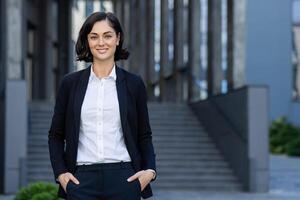 profesional mujer de negocios en un inteligente traje en pie con confianza fuera de un oficina edificio en un urbano configuración, retratar éxito y liderazgo. foto