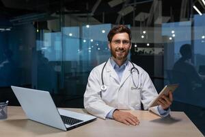 retrato de exitoso maduro médico en médico Saco con estetoscopio barba y anteojos, hombre participación tableta computadora trabajando sentado a mesa con computadora portátil, sonriente y mirando a cámara. foto