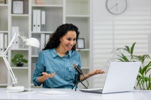 Cheerful professional woman recording a podcast in a contemporary home office set up with a laptop and microphone. photo