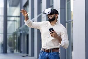 Photo of a young Muslim man in a shirt standing outside an office building wearing a virtual mask, holding a phone, talking online and gesturing with his hands