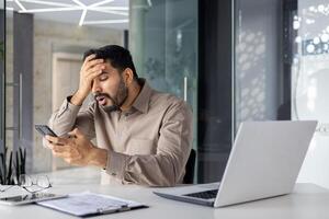 un indio empresario mirando confuso y estresado como él examina su teléfono inteligente en un moderno oficina configuración, con un ordenador portátil y documentos en el escritorio. foto