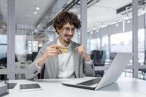 A youthful male entrepreneur with curly hair, beaming as he confidently holds a credit card in a modern office setting. photo