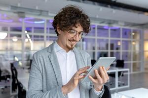 Content young male office worker using a digital tablet, the epitome of professional ease and technological proficiency in a bright office setting. photo