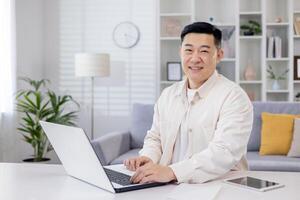 retrato de exitoso asiático programador, hombre trabajando remotamente desde hogar oficina, empresario sonriente y mirando a cámara utilizando ordenador portátil a trabajo sentado en vivo habitación a hogar. foto