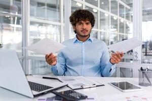 Portrait of angry disappointed financier inside office at workplace, hispanic businessman holding contracts reports in hands and looking nervously at camera, upset man behind paper work. photo