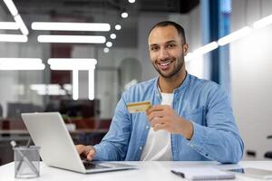 un sonriente hombre participación un crédito tarjeta mientras utilizando un computadora portátil, representando seguro en línea actas y digital bancario. foto