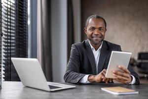 A professional African American businessman engages with a digital tablet at his desk in a modern office setting, portraying confidence and expertise. photo