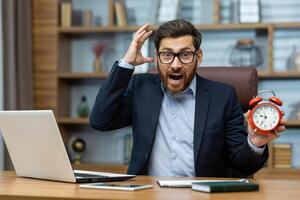 Surprised mature businessman in a suit holds a red alarm clock, expressing urgency, deadline stress in home office. photo