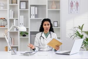 A professional female doctor in a white lab coat sits at her office desk, appearing concerned while holding medical records and discussing healthcare issues. photo