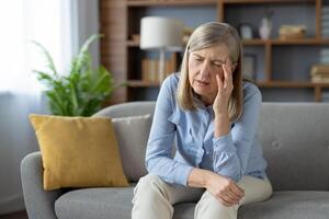 An elderly woman appears distressed and alone, sitting pensively on a couch in a well-lit living room, evoking emotions of loneliness and concern. photo