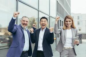 Interracial business partnership. Three businessmen celebrate a victory, a successful deal. An older man, a young asian man and a young woman in suits near office, laugh, show with their hands yes. photo