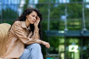 Worried young woman sitting outdoors, holding glasses, deep in thought with a troubled expression, urban backdrop. photo