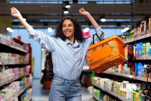 retrato de un contento mujer comprador en un supermercado, un Hispano mujer con un cesta de bienes sonrisas con Placer y bailes entre el estantería con bienes, un satisfecho tienda cliente foto