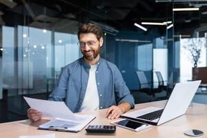 exitoso financiero inversor trabajos con documentos y cuentas dentro oficina, empresario con barba sonriente satisfecho con trabajo resultados y logros, hombre dentro oficina con ordenador portátil laboral. foto