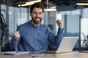 un alegre empresario celebra un exitoso acuerdo o logro mientras trabajando en su ordenador portátil a el oficina. foto