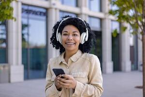 Portrait of young beautiful African American woman, student smiling and looking at camera holding phone in hands using headphones for listening to music and online radio with podcasts. photo