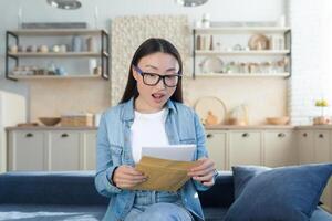 Happy asian girl shocked by received letter, woman reading message sitting on sofa at home photo