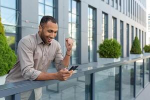 A happy Hispanic businessman standing near the office. Holding phone, playing online games photo