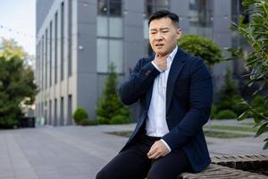 Asian young male businessman sitting on a bench on the street near an office building and holding his hand to his throat, feeling severe pain and discomfort. photo