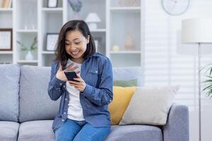 mujer celebrando victoria a hogar mirando a teléfono pantalla, satisfecho asiático mujer contento recibido en línea notificación de bueno logro resultados sentado en sofá en vivo habitación. foto