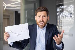 A business professional expressing frustration and confusion while holding a graph showing stock market fluctuations in an office setting. photo