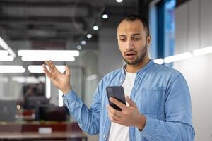 Indian man in casual attire expressing surprise while looking at his smartphone in a contemporary office setting. photo