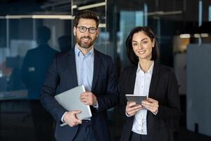 sonriente masculino y hembra en elegante trajes en pie juntos en oficina salón y participación digital artilugio en manos. simpático compañeros de trabajo consiguiendo Listo para en línea reunión con jefe siguiente a sala del consejo. foto