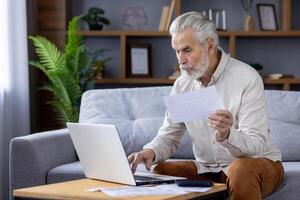 Focused senior man with grey hair, working on a laptop while sitting on a couch, attentively reviewing documents in a cozy home environment. photo