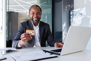 Bank employee, smiling young African American man sitting in office at desk with laptop, holding and showing credit card to camera. photo