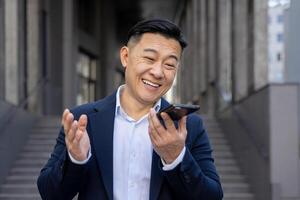 Happy Asian businessman in blue suit engaging in a light-hearted conversation on his phone, laughing joyously outside. photo