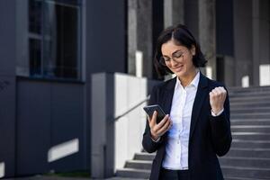 joven hermosa negocio mujer en un negocio traje caminando en el ciudad con un teléfono en manos, un mujer usos un solicitud recibido un notificación de un victoria ganar, celebra triunfo y logro. foto