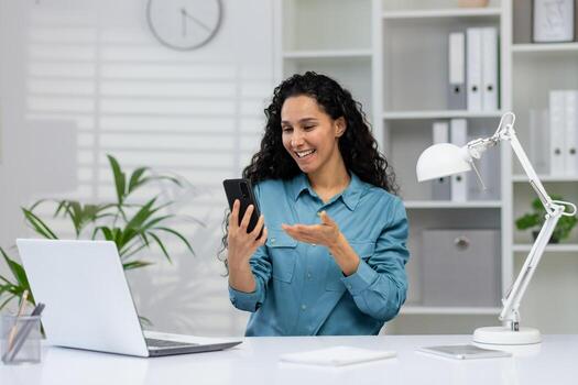 un sonriente, profesional mujer se involucra en un llamar. ella es sentado a un limpio, organizado oficina escritorio, gesticulando mientras mirando a su teléfono inteligente foto