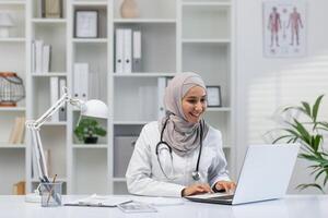 positivo y simpático musulmán hembra médico disfrutando su trabajo en un brillante oficina ambiente, revisando médico datos en ordenador portátil. foto