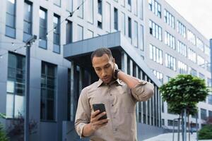 Pensive and excited african american worker outside office building reading bad news, man holding phone photo